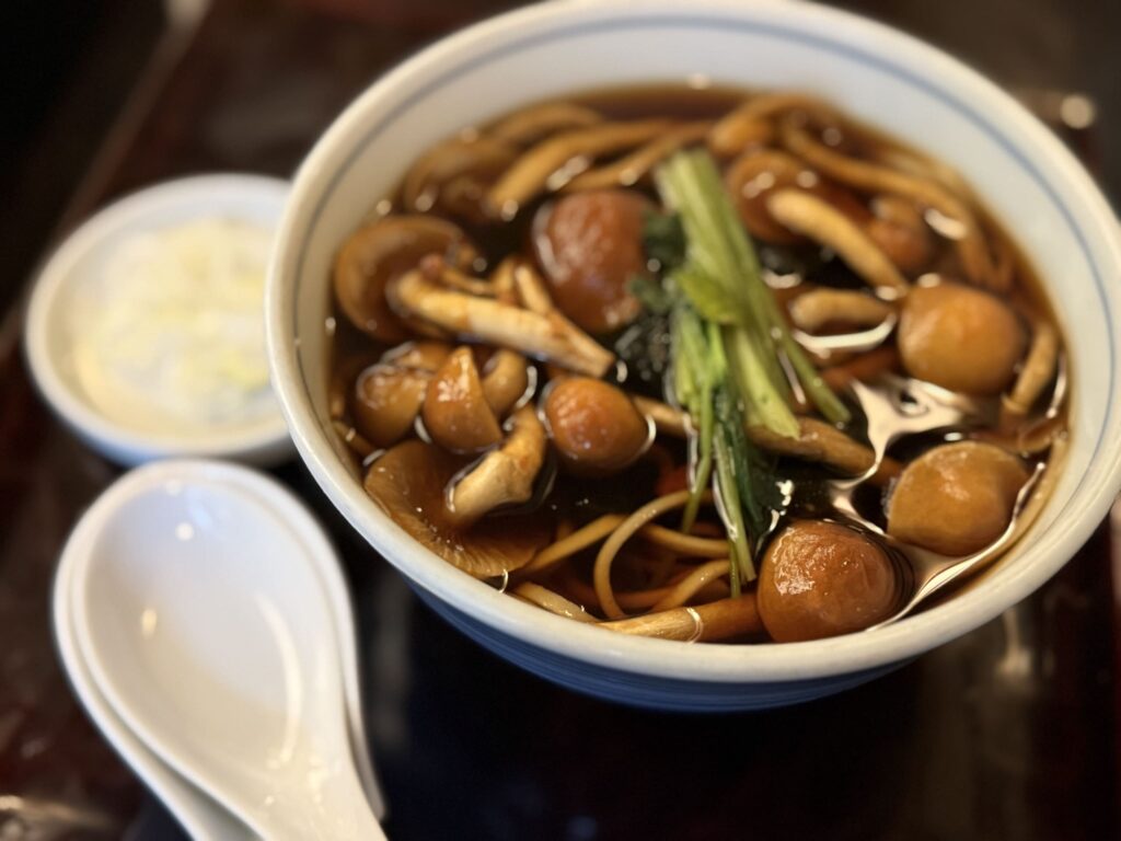 A close-up of a bowl of hot soba noodles filled with various mushrooms, such as nameko and shimeji, placed on a tray. A small bowl with grated yam and a white ceramic spoon are placed beside it.