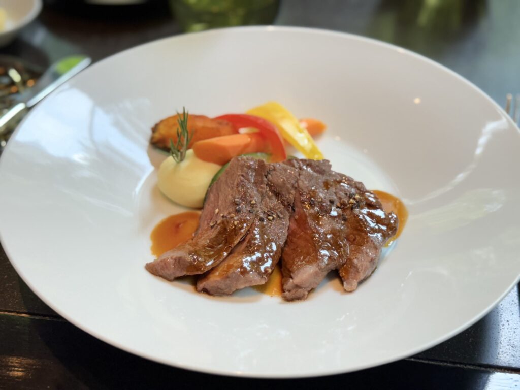 A close-up of grilled Japanese beef loin served on a white plate. The sliced meat is covered in a glossy sauce and accompanied by mashed potatoes and colorful cooked vegetables like carrots and bell peppers.
