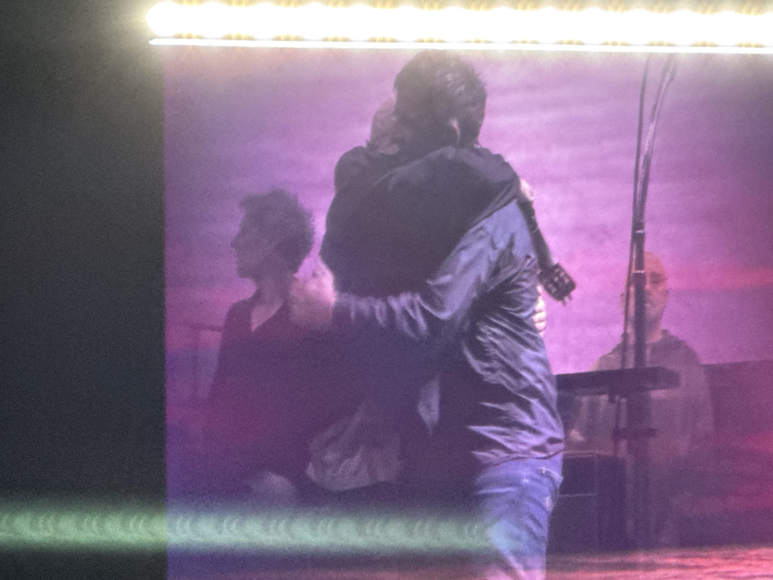 A person holding a guitar and another person embracing tightly on a stage bathed in purple light.