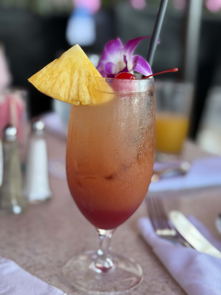 A tropical cocktail in a glass on a restaurant table, garnished with a slice of pineapple, a cherry, and a purple orchid flower.