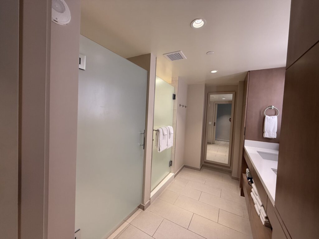 A spacious hotel bathroom. There is a frosted glass door in the foreground on the left, a shower stall further back, and a vanity on the right.