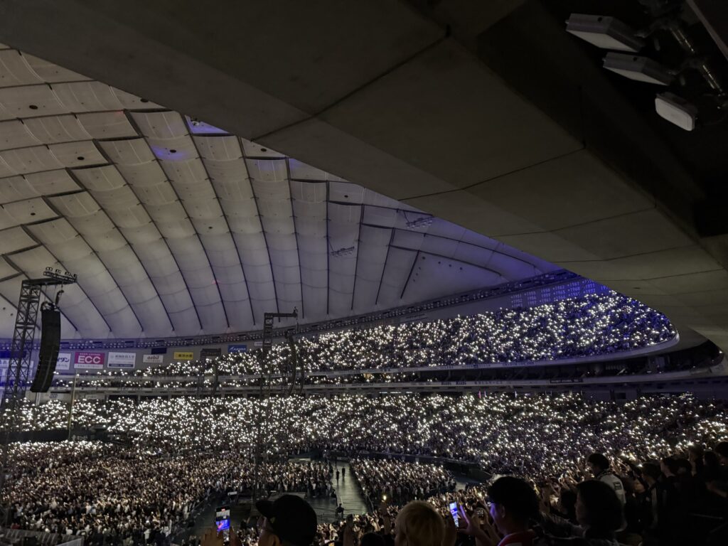 A view from the stands of a large dome concert venue, where the audience is holding up their smartphone flashlights, creating a sea of bright white lights.