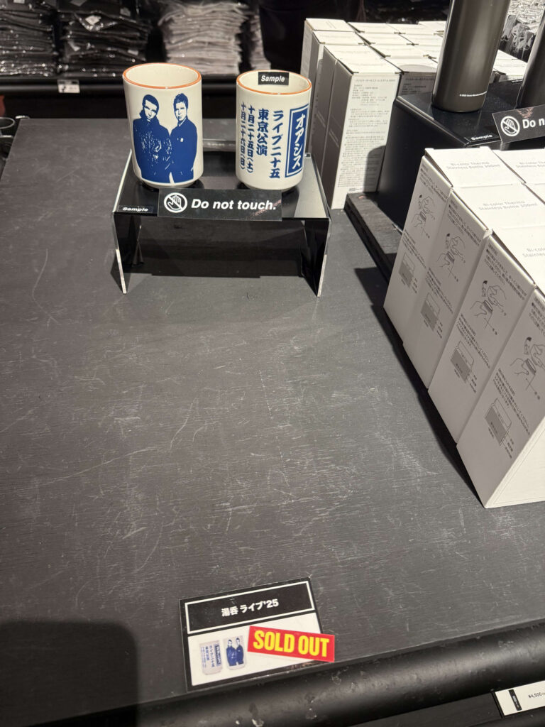 A sample of Oasis commemorative Japanese-style teacups (yunomi) on a display stand. In the foreground, the item tag has a red 'SOLD OUT' sticker on it.