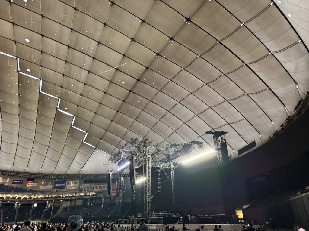 A dome stadium before a concert. A large stage is set up in the arena, with audience seats spreading out under the characteristic white panelled ceiling.