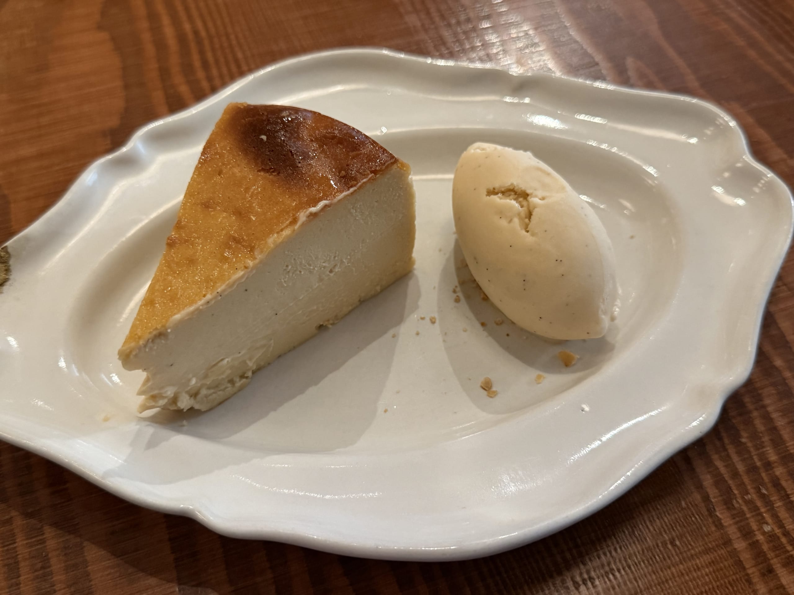 A slice of Basque cheesecake with a scoop of vanilla bean ice cream on a white, wavy plate, placed on a wooden table.