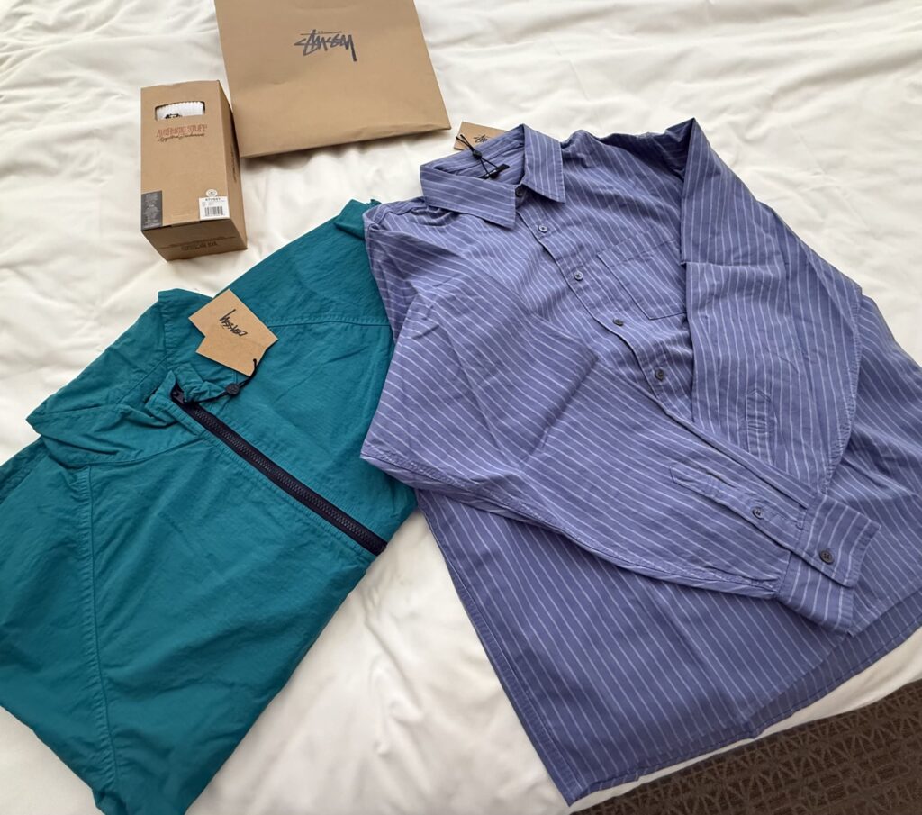 A teal Stussy warm-up jacket and a blue striped shirt are spread out on a white sheet. A Stussy paper bag and a box are placed in the background.