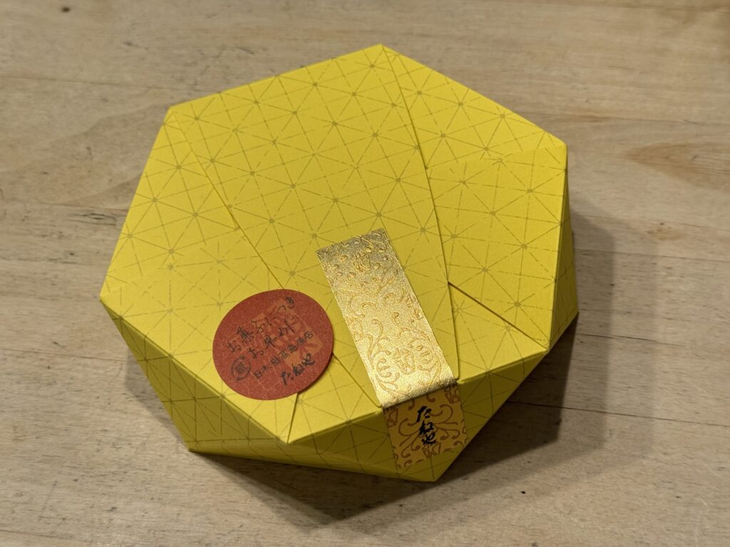 木製のテーブルの上に置かれた、たねやの和菓子「きぬかづき」が入った黄色い八角形の箱。幾何学模様がデザインされており、金色の帯と赤い丸いシールで封がされている。