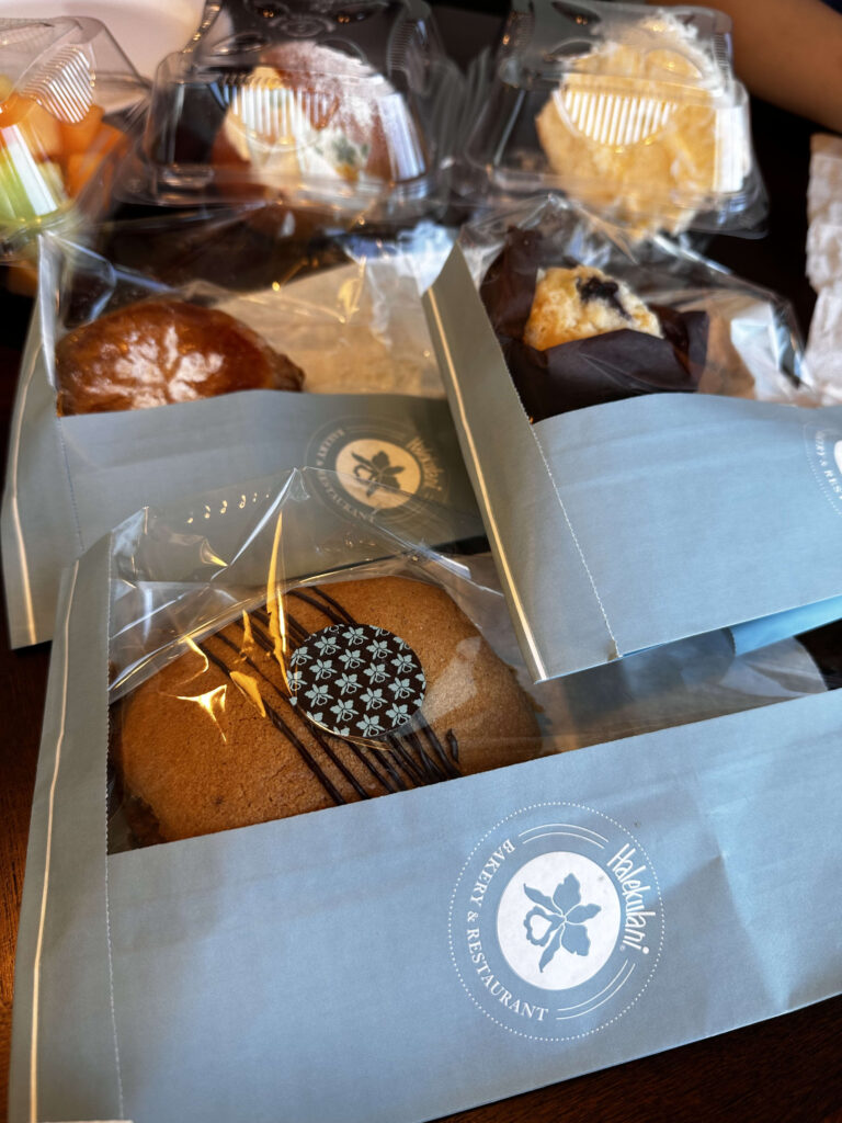 A collection of individually wrapped pastries, including a cookie and a muffin, in light blue paper bags from Halekulani Bakery & Restaurant.