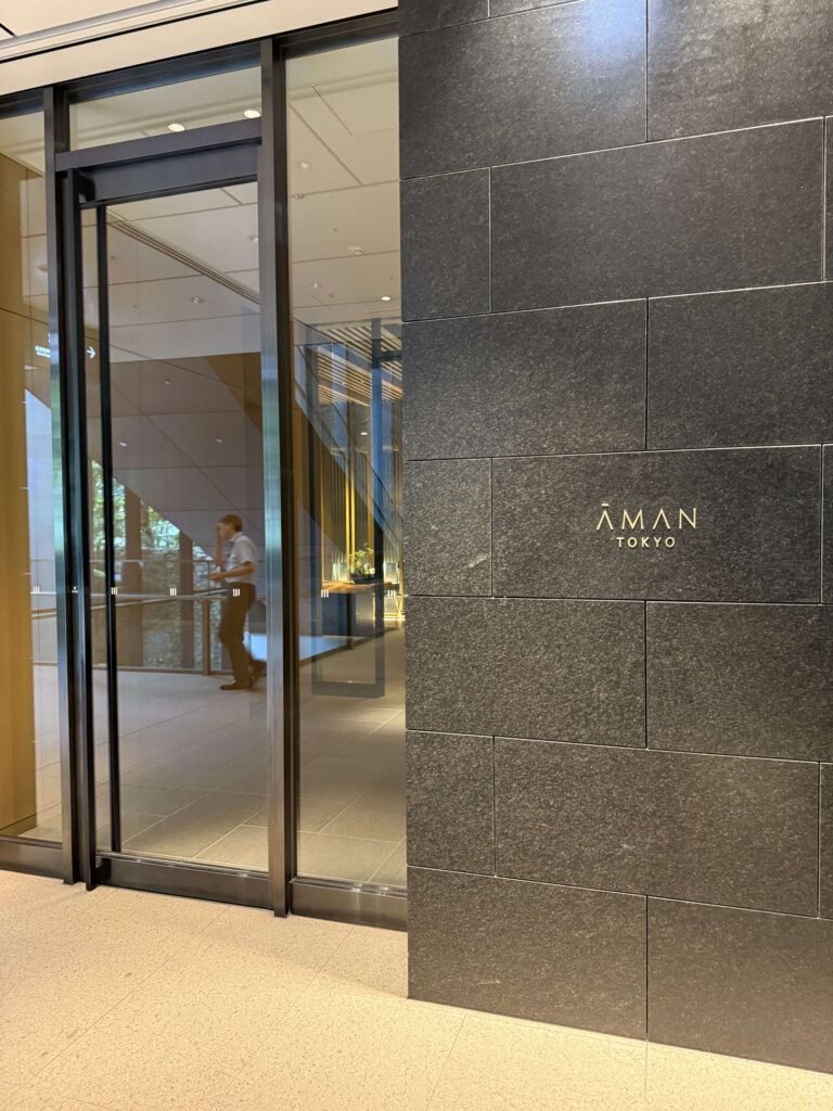 アマン東京のエントランス。黒い石壁に金色のロゴが刻まれ、ガラス張りの自動ドアの向こうにモダンな内装が見える。