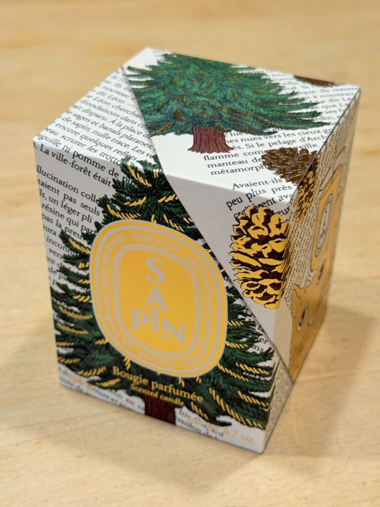 The box for the Diptyque limited edition holiday candle 'Sapin' on a wood-grain table. The design features illustrations of fir trees and pine cones.