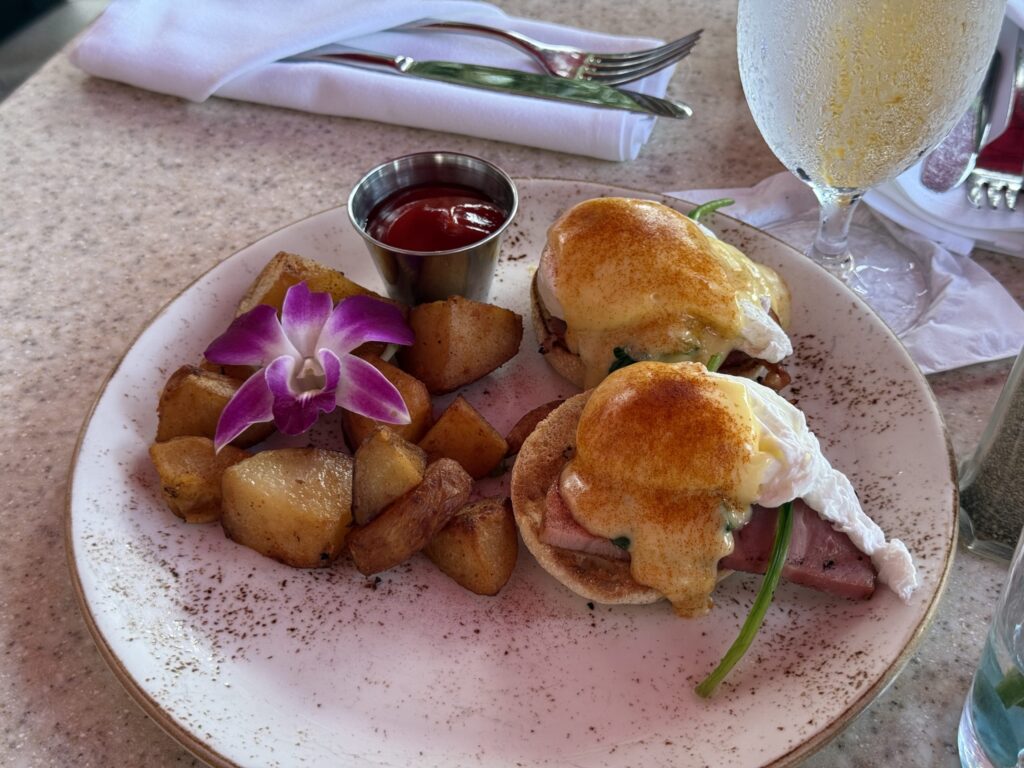 A plate of Eggs Benedict with a side of roasted potatoes. Two poached eggs are covered in a creamy hollandaise sauce, and the dish is garnished with a purple orchid flower.