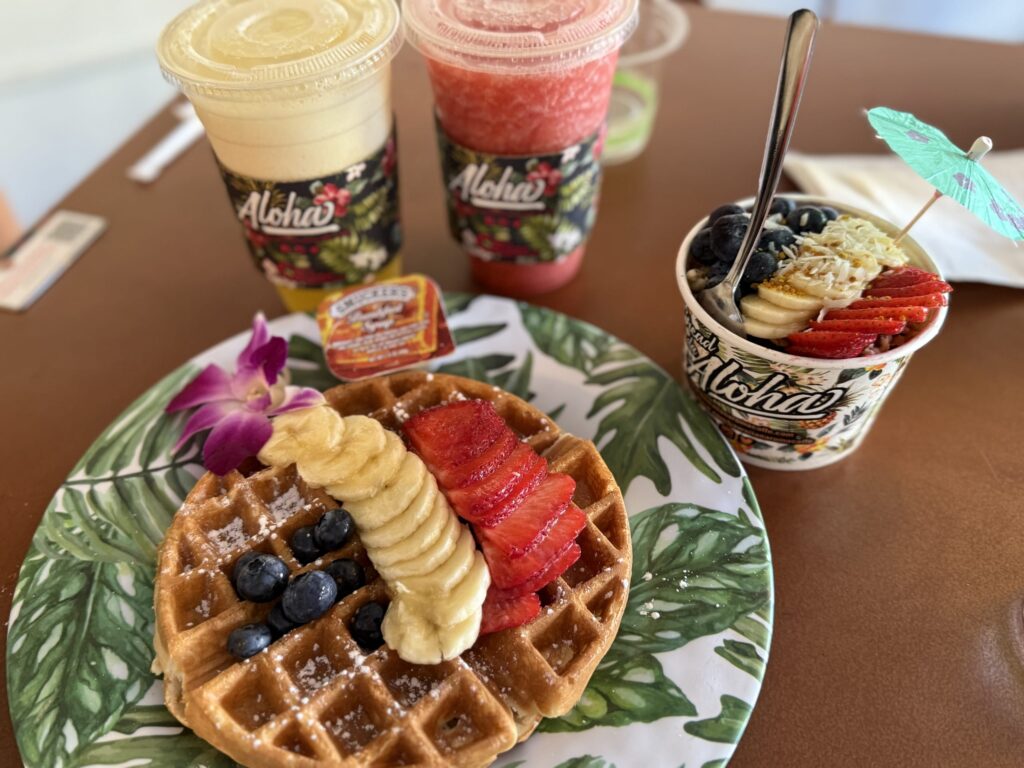 A fruit-topped waffle, an acai bowl, and two kinds of smoothies placed on a table.