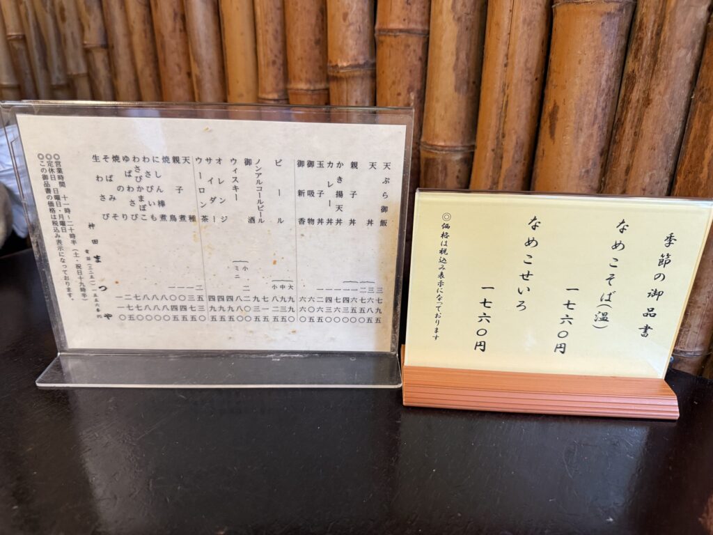 The menu of the long-established soba restaurant 'Kanda Matsuya' placed on a table against a bamboo wall background, showing a detailed menu and a seasonal menu for 'Nameko Soba'.