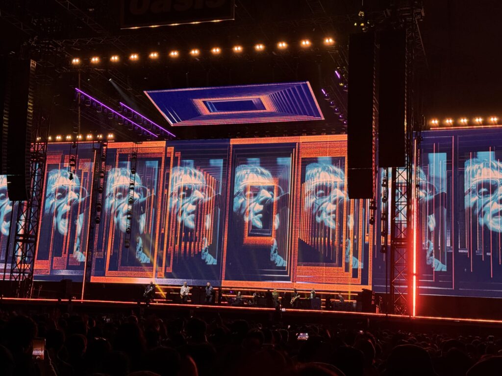 The stage at an Oasis live concert. Giant LED screens display a repeating, psychedelic pattern of Liam Gallagher's face in sunglasses with orange and blue effects. The band performs on stage as the venue is filled with a large audience.