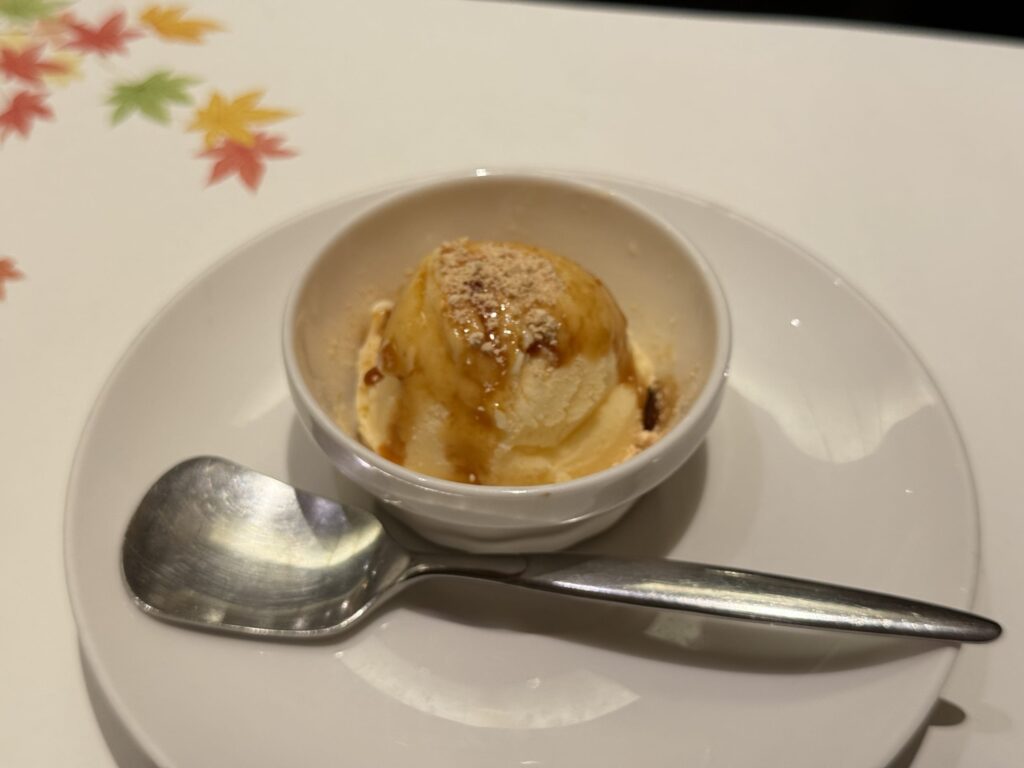 A scoop of soy milk ice cream topped with kinako powder and kuromitsu syrup in a white bowl. A colorful maple leaf pattern is visible in the background.