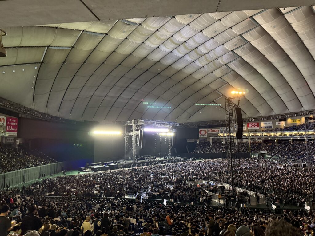 ドーム球場のスタンド席から撮影されたコンサート会場の全景。中央には明るく照明が当てられたステージが設置され、アリーナとスタンドは開演を待つ大勢の観客で埋め尽くされている。