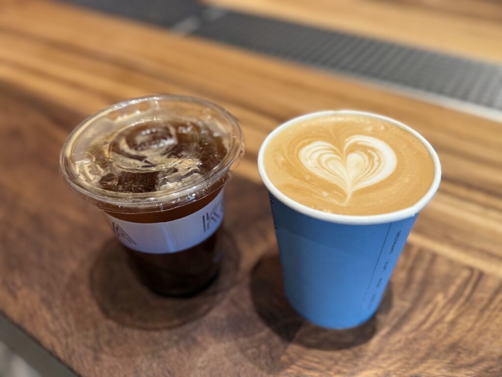 An iced Americano in a plastic cup and a Caffe Latte in a blue paper cup with heart-shaped latte art, placed on a wooden counter.