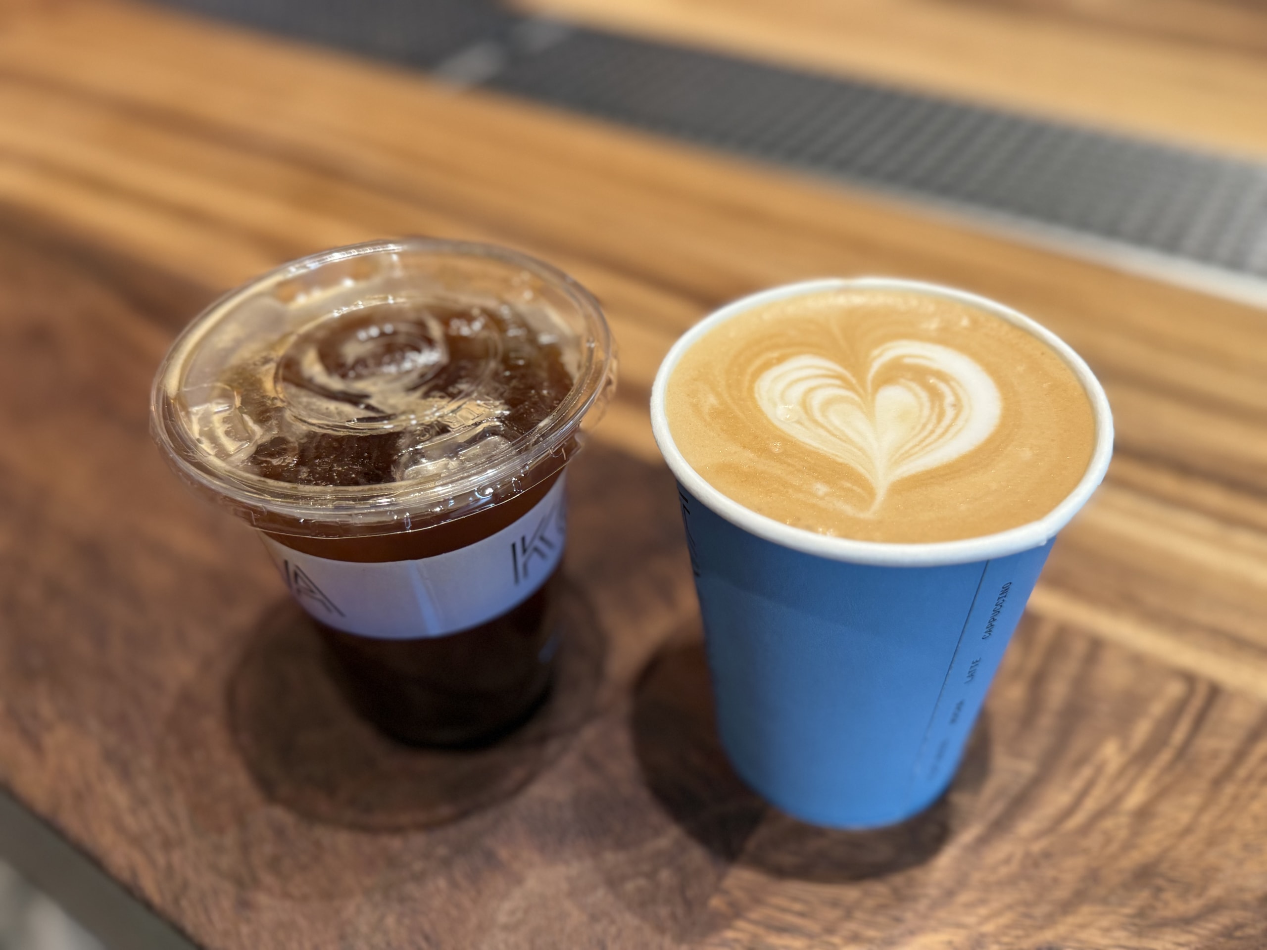 An iced Americano in a plastic cup and a Caffe Latte in a blue paper cup with heart-shaped latte art, placed on a wooden counter.