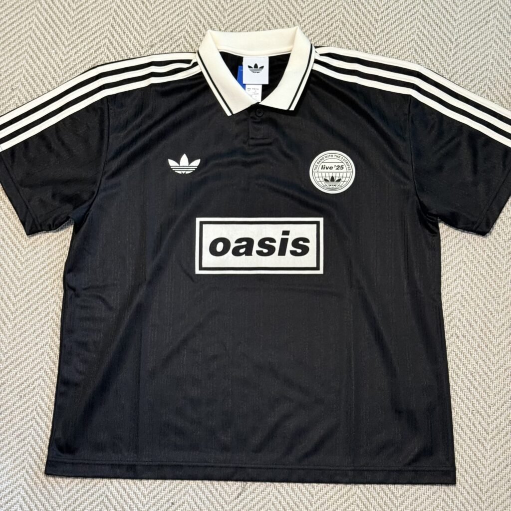 A black Adidas x Oasis collaboration game shirt laid flat on a herringbone patterned surface. It features the Oasis box logo on the chest, three stripes on the shoulders, and a 'live '25' patch on the left breast.