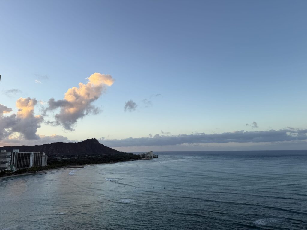 高層階から見た早朝のワイキキビーチとダイヤモンドヘッド。穏やかな海と海岸線に並ぶホテル群、そして朝日を浴びてピンク色に染まる雲が空に浮かんでいる。
