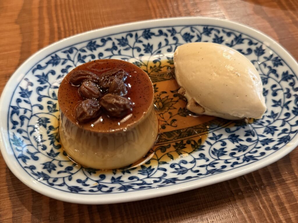 A custard pudding with rum raisins and a scoop of vanilla ice cream on a blue and white patterned plate, placed on a wooden table.
