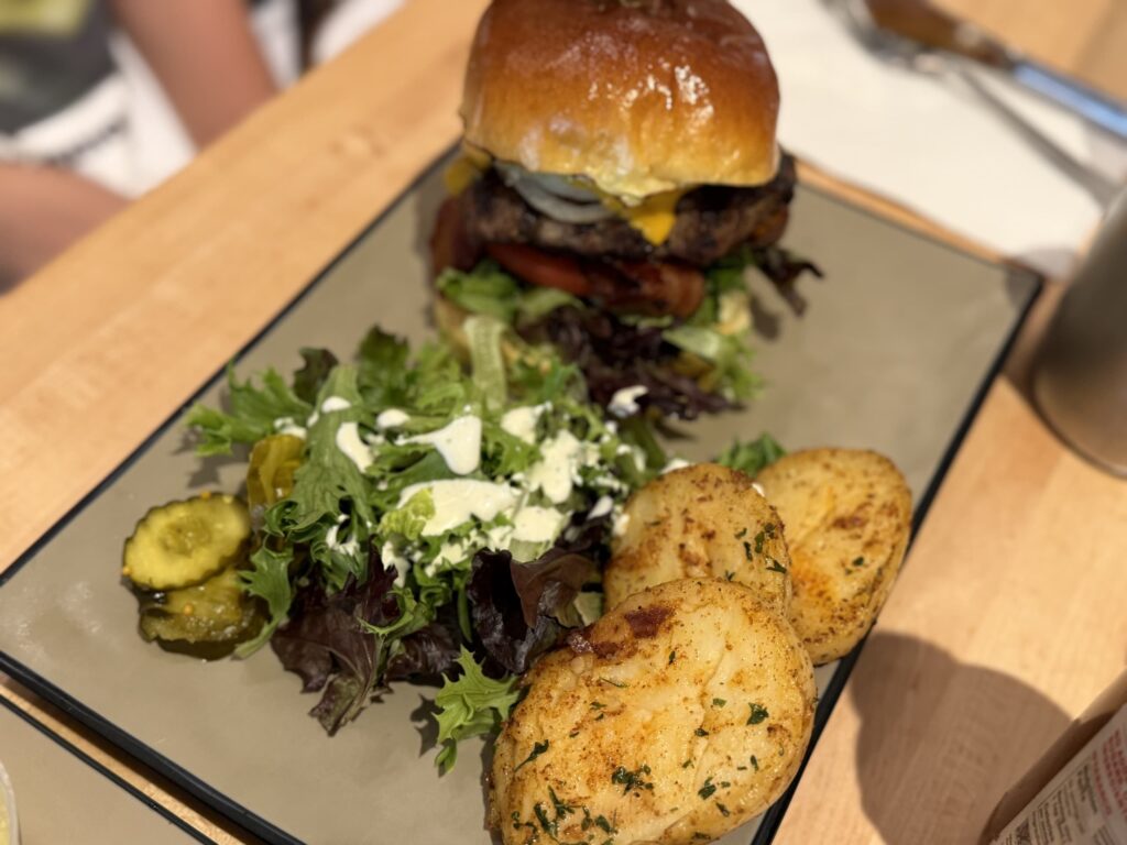 木のテーブルの上に置かれた四角い皿。皿の上には、大きなチーズバーガー、ローストポテト、クリーミーなドレッシングがかかったグリーンサラダ、ピクルスが盛り付けられている。