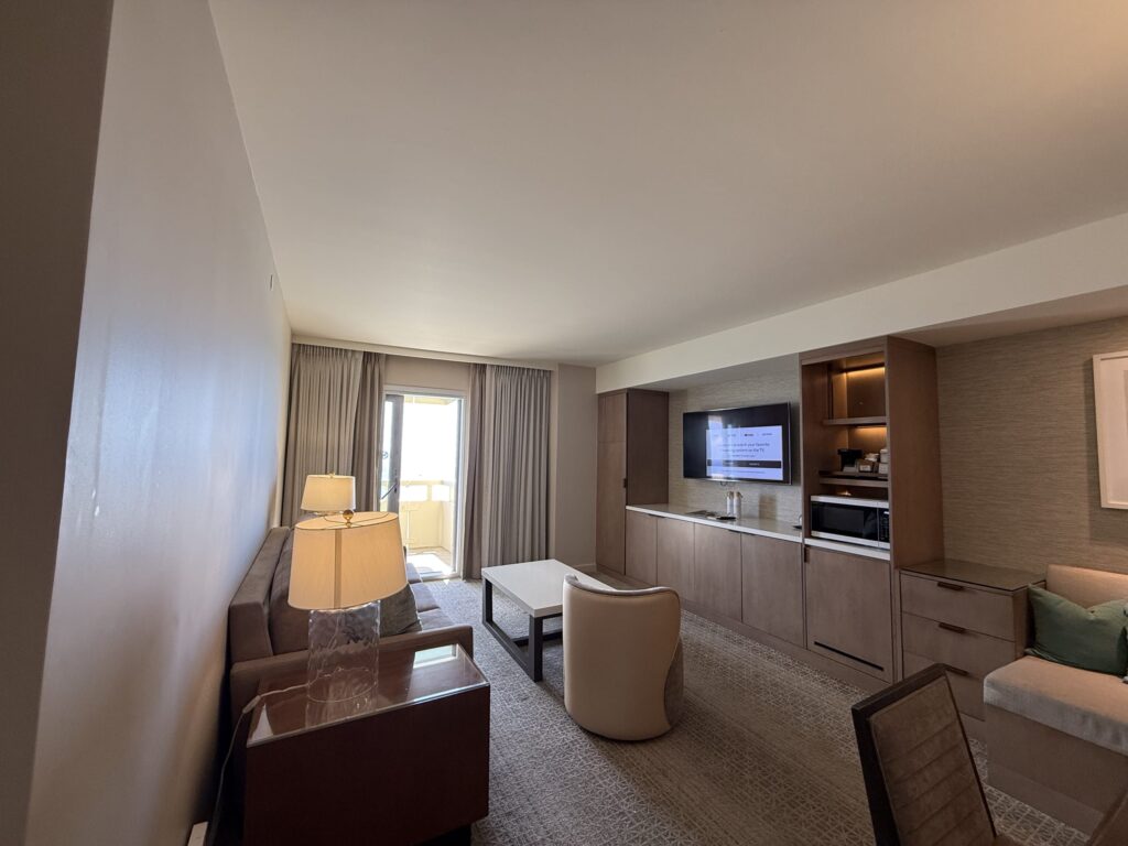 A spacious hotel living room with a sofa, TV, coffee table, and a balcony visible in the background.