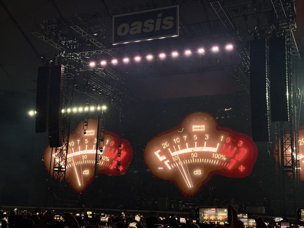 暗いコンサート会場のステージ。上部に「Oasis」のロゴが掲げられ、2つの巨大なVUメーターのグラフィックがスクリーンに赤く光っている。下には多くの観客がスマートフォンを掲げて撮影している。