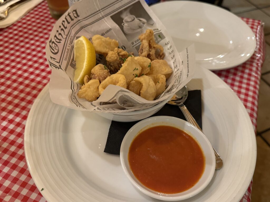 A white plate on a red checkered tablecloth, holding a serving of fried calamari in a bowl lined with newspaper-style paper, accompanied by a lemon wedge and a small bowl of red dipping sauce.