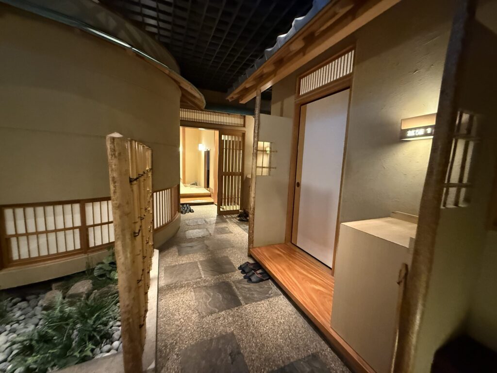 A corridor in a traditional Japanese style. The path is paved with flagstones and gravel, and lined with entrances to private rooms featuring wooden doors and shoji paper screens.