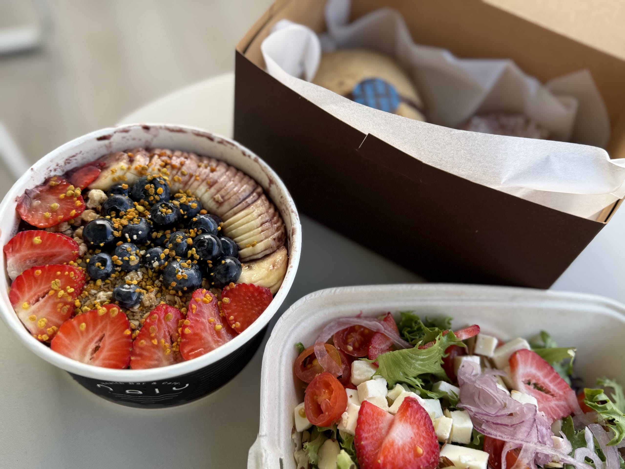 An acai bowl topped with fresh fruit and a salad in takeout containers on a white table, with a pastry box in the background.