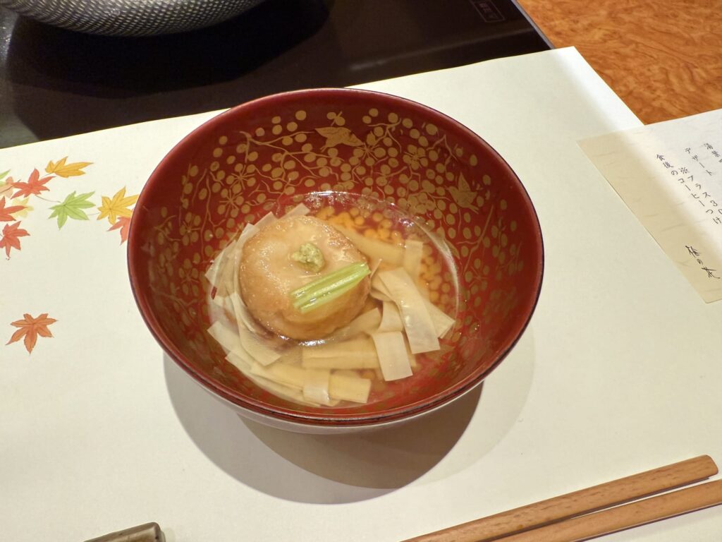 A fried lotus root dumpling in a red bowl with gold patterns. It is soaking in a clear broth and garnished with wasabi. A paper with autumn leaf illustrations is visible on the left.