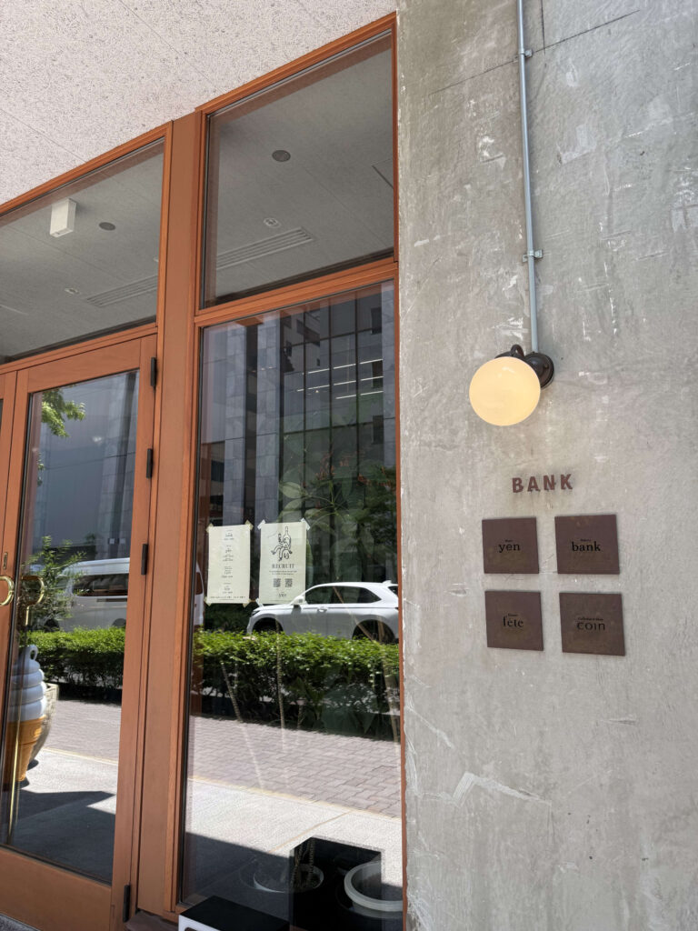 The entrance to the 'BANK' complex, featuring a concrete wall and wooden-framed glass doors. Four signs for the shops 'yen,' 'bank,' 'fete,' and 'coin,' along with a round lamp, are mounted on the wall.