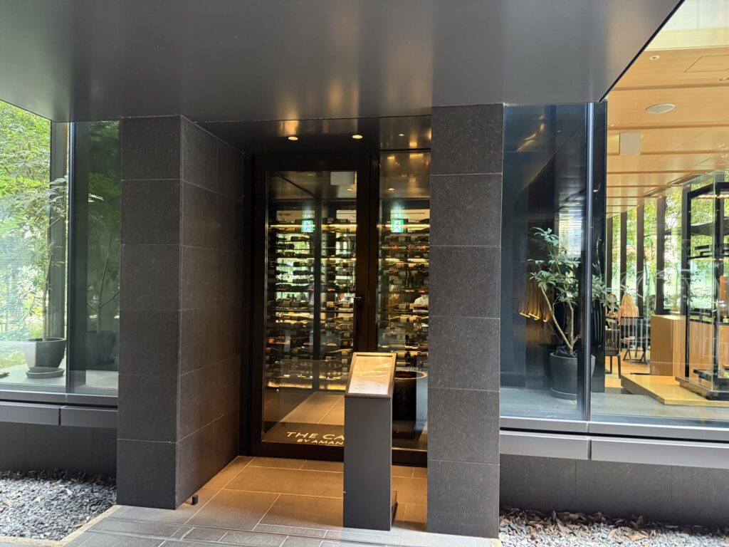 The modern entrance to a cafe, featuring glass doors surrounded by black stone walls. A wine cellar is visible inside.