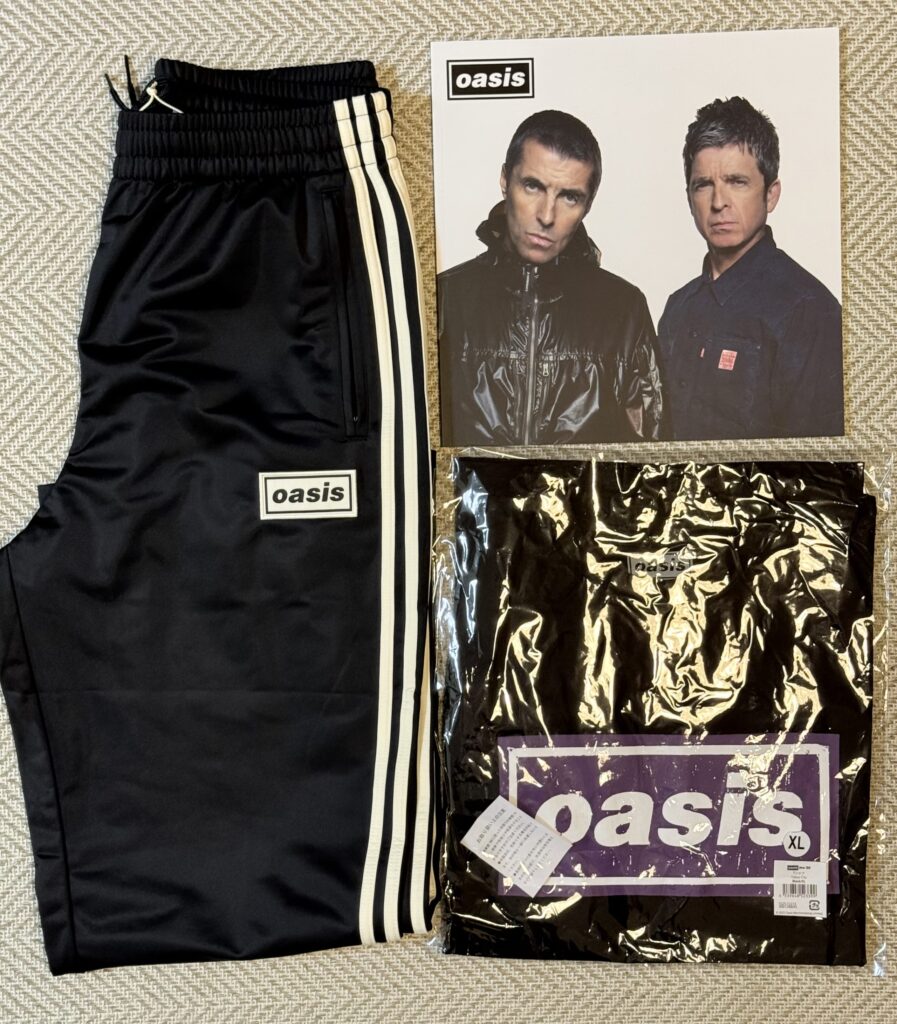 ベージュの布地の上に置かれたOasisのグッズ。左から、Oasisのロゴが入った黒いトラックパンツ、リアムとノエル・ギャラガーが写ったツアーパンフレット、パッケージに入った黒いTシャツ。