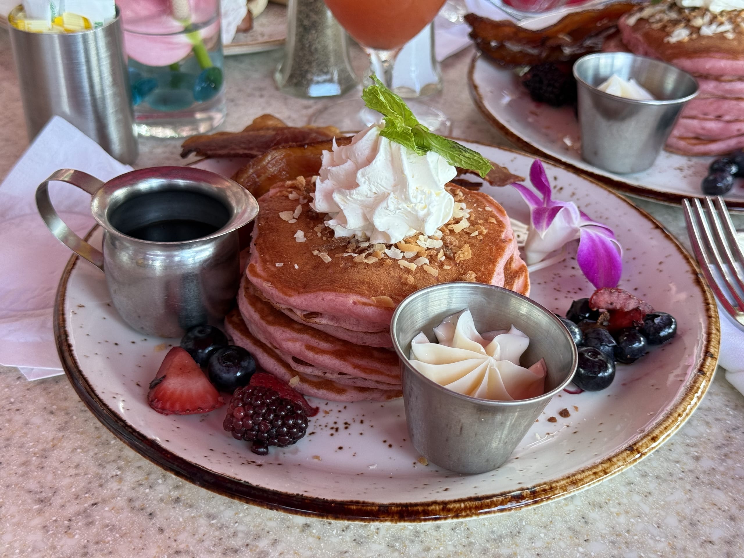 A stack of pink pancakes topped with whipped cream, toasted coconut flakes, and a mint sprig, served with fresh berries, bacon, syrup, and butter.