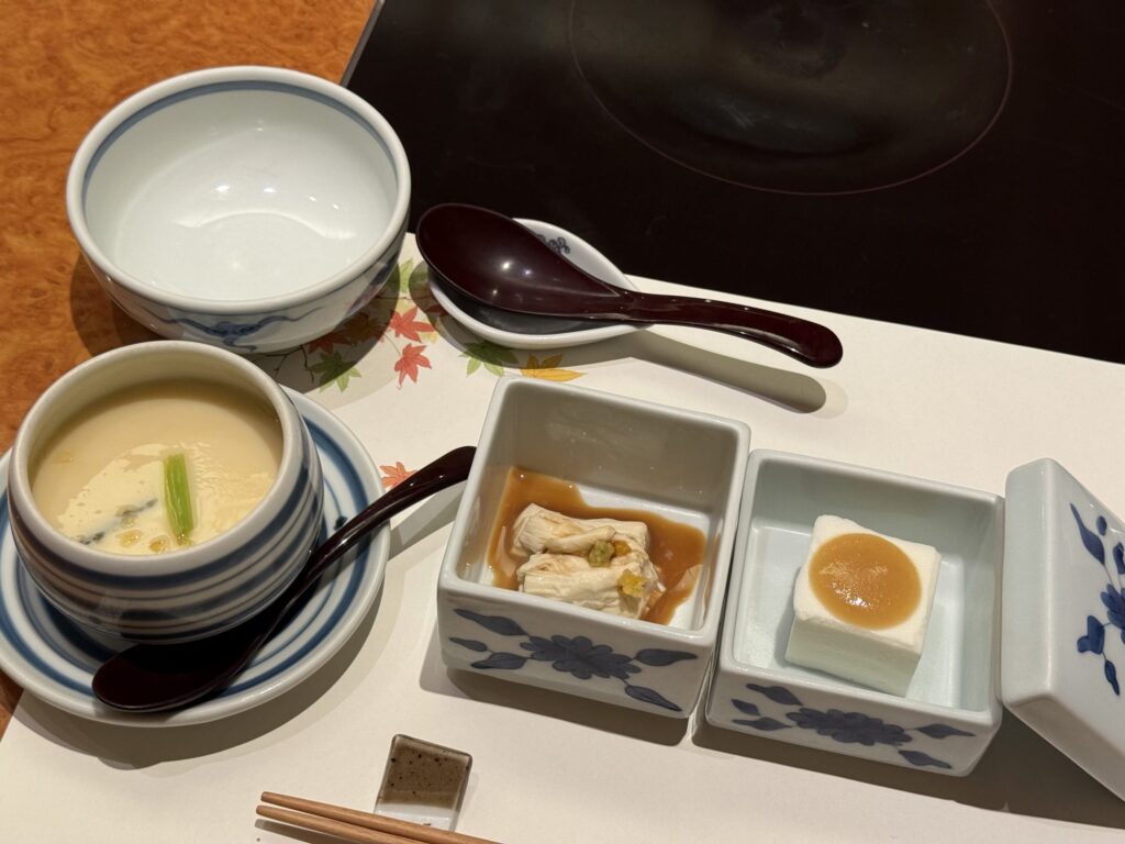 A Japanese meal set on a table, featuring chawanmushi in a blue and white striped bowl, and two square dishes containing yuba sashimi and soy milk tofu.