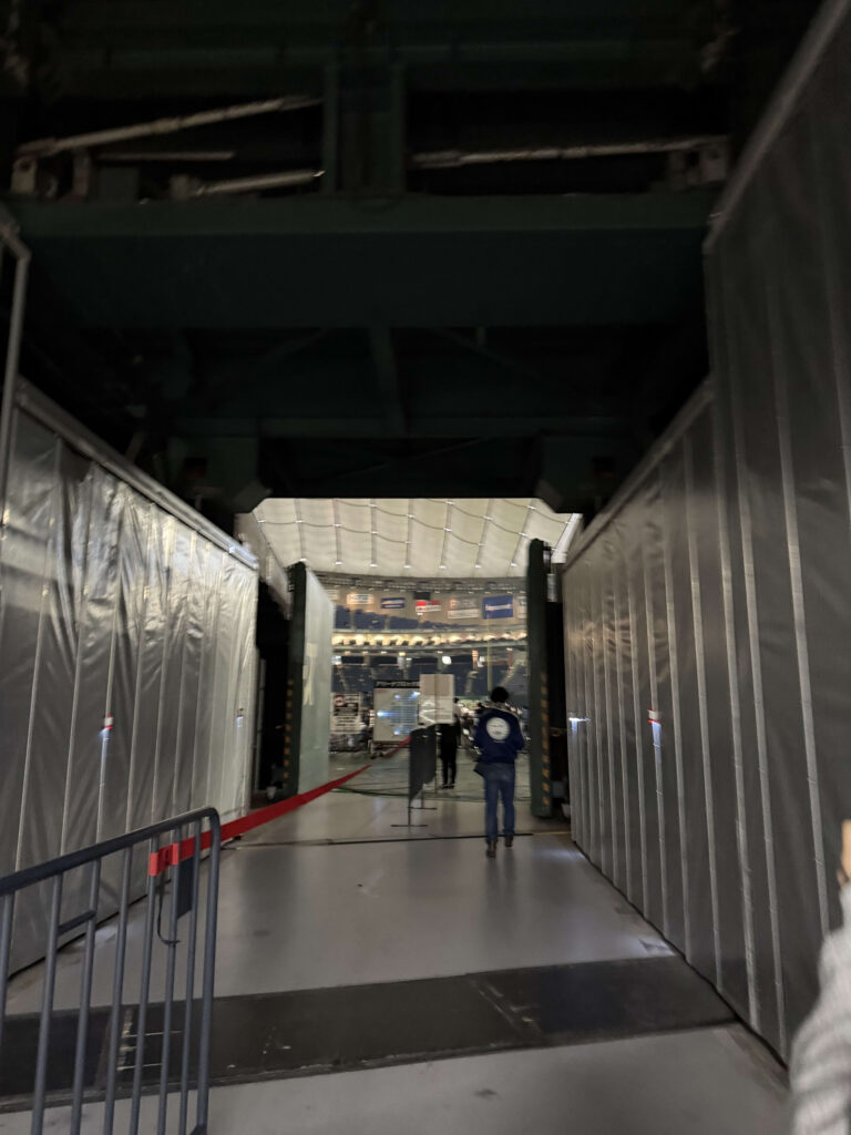 View from a dark backstage corridor looking out into a brightly lit arena. A person is seen from behind walking down the passage toward the light.