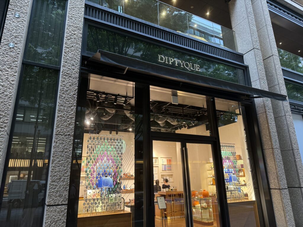 夕暮れ時のdiptyqueブティックの外観。石造りの柱と大きなガラス窓が特徴で、温かい光に照らされた店内とカラフルなディスプレイが見える。