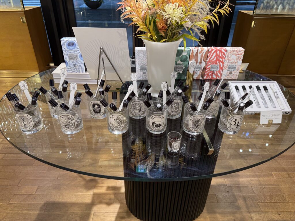 A round glass table in a Diptyque store displaying various fragrance testers in glass tumblers, a selectable discovery set, and a vase of autumn flowers in the background.