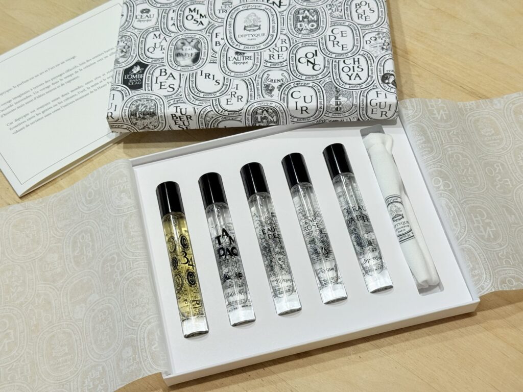 木製のテーブルの上に置かれたディプティックの香水セット。開いた白い箱の中に、5本のトラベルサイズ香水スプレーと白い布製ポーチが並んでいる。