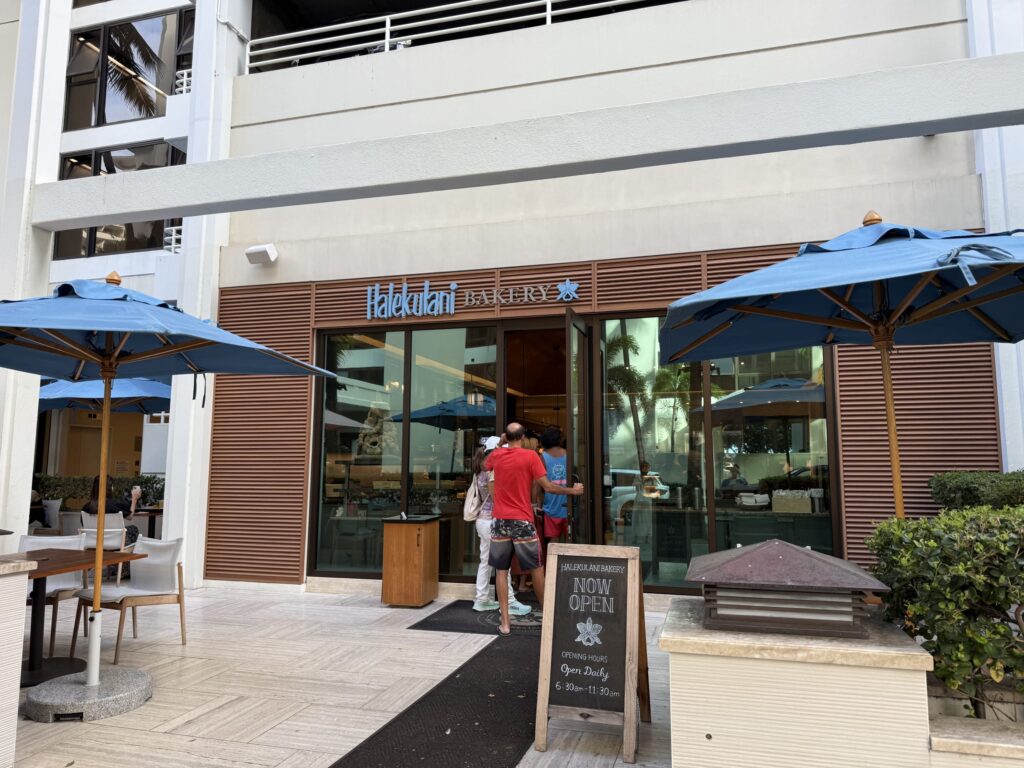 The modern exterior of Halekulani Bakery. Several customers are lined up at the entrance waiting to get in, and there are patio tables with blue umbrellas in the foreground.