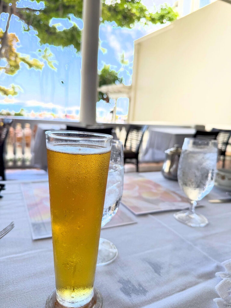 A glass of cold beer with condensation on it, placed on a table at a restaurant terrace. A bright, sunny scene spreads out in the background.