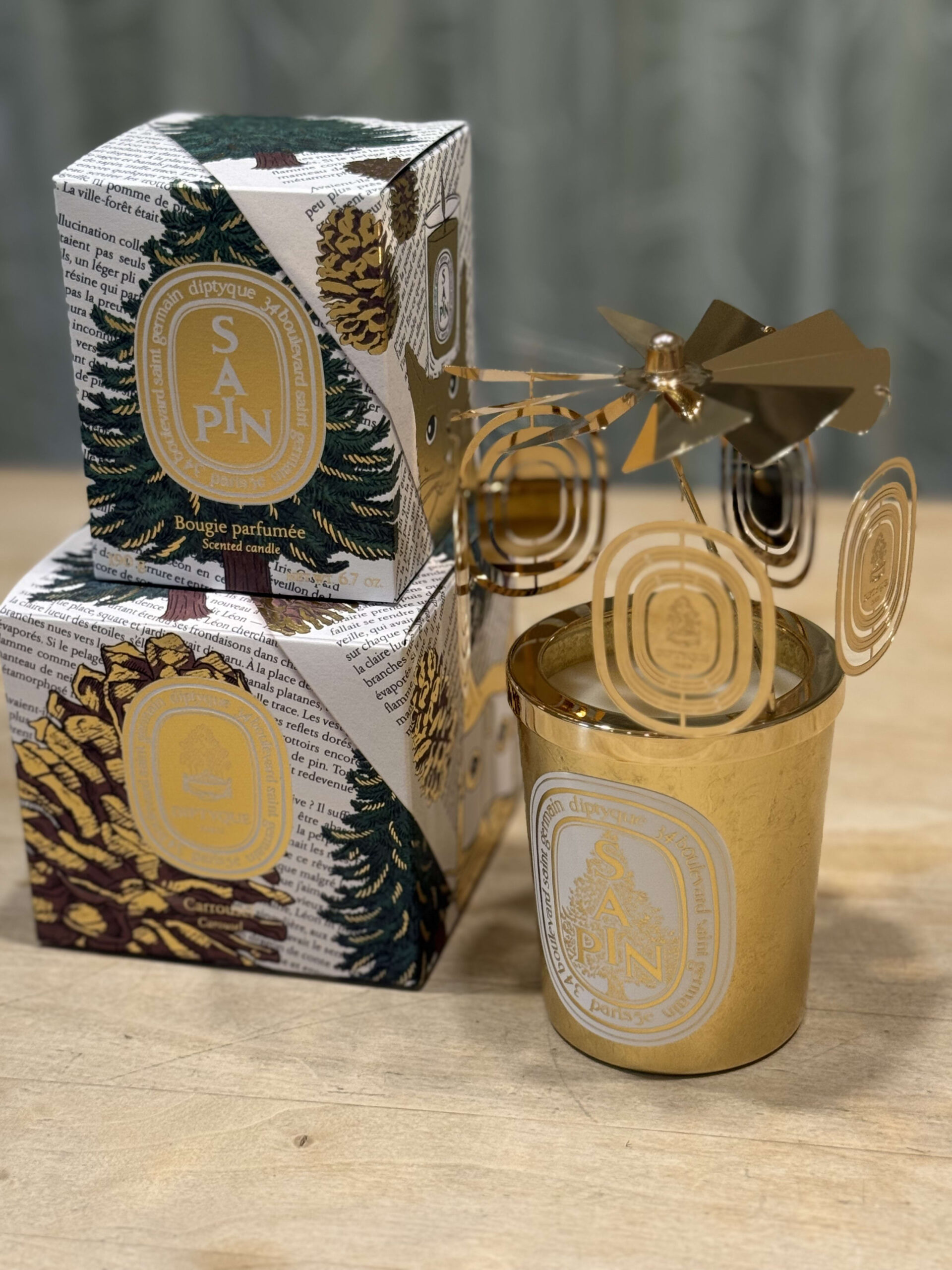 The Diptyque holiday collection on a wooden table. A golden carousel is placed on top of the 'Sapin' candle in its gold vessel, with two stacked gift boxes illustrated with pinecones and fir trees next to it.