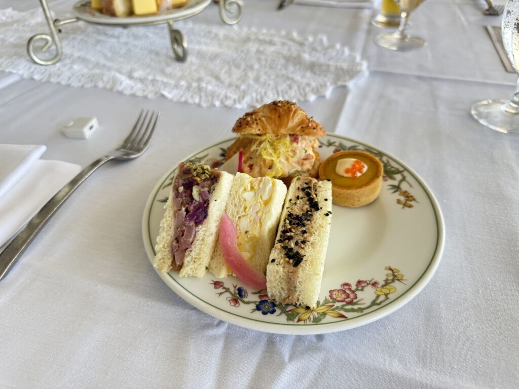 A plate of afternoon tea savories on a floral plate, placed on a white tablecloth. The plate holds several types of finger sandwiches, a mini croissant sandwich, and a small tart.