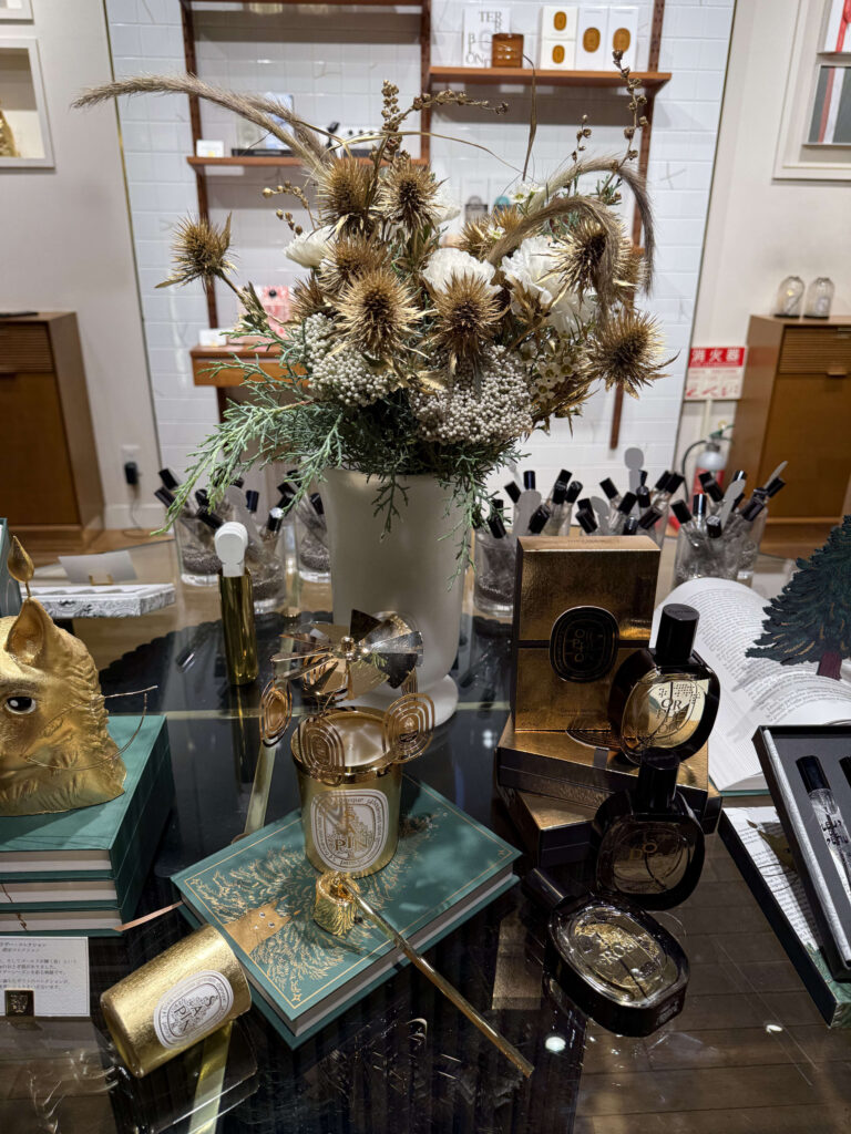 A holiday collection display at the diptyque Marunouchi store, featuring limited edition candles and perfumes arranged around a centerpiece of gold and white dried flowers.