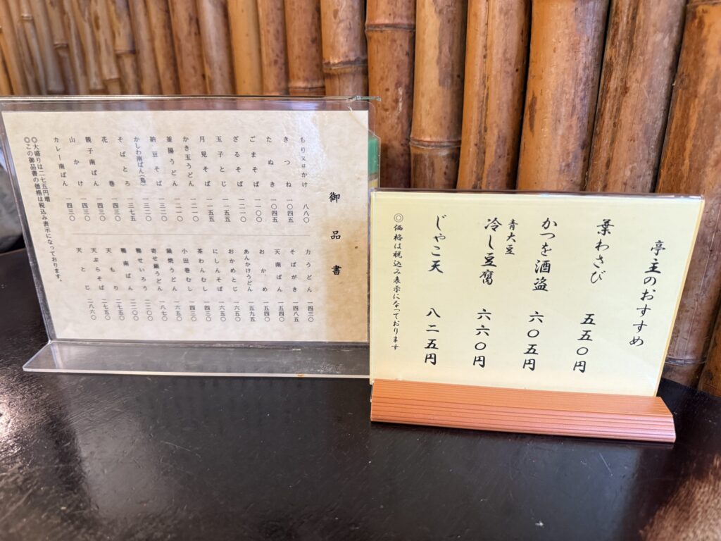 Two types of menus on a table at the long-established soba restaurant Kanda Matsuya. The main menu is on the left, and a vertical menu with the 'Owner's Recommendations' is on the right, with a bamboo wall in the background.