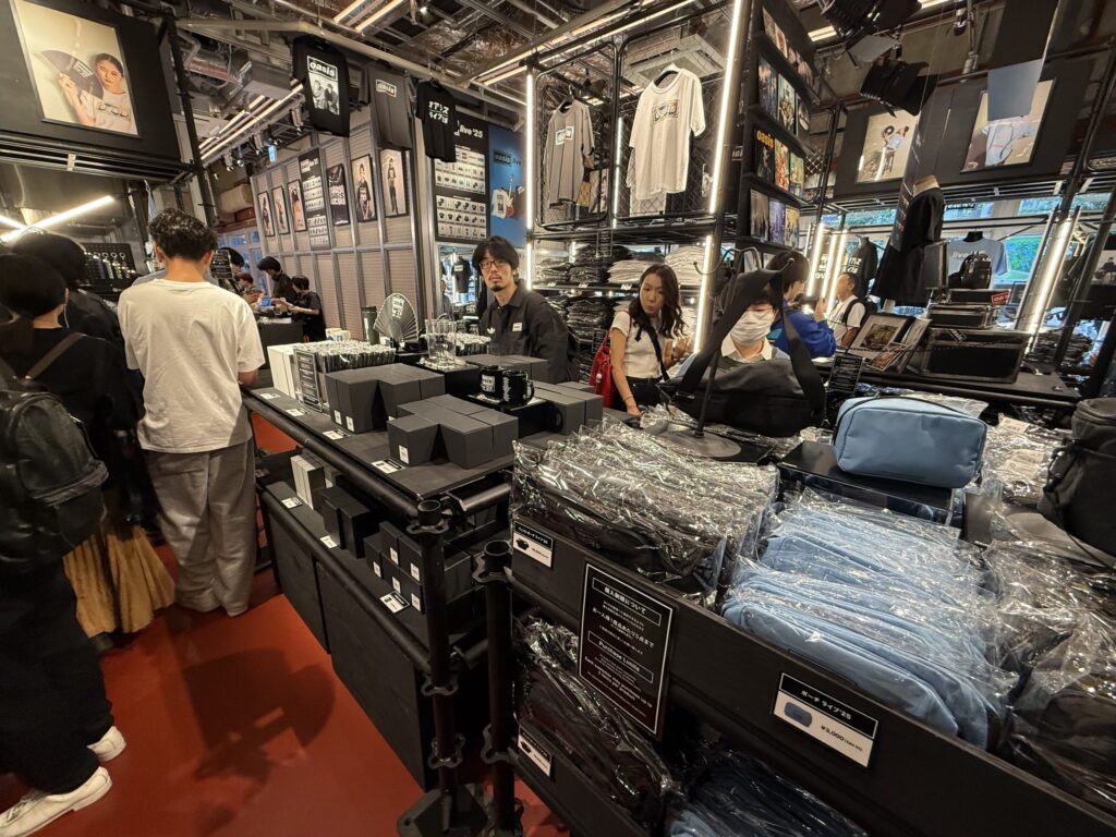 オアシスのポップアップストアの店内。多くの客がレジに並び、Tシャツやマグカップなどのグッズが陳列されている。