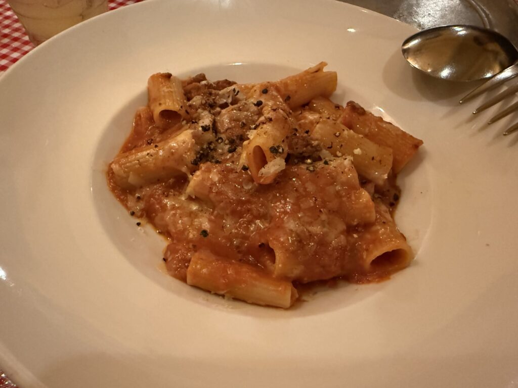 A white plate of beef meat sauce pasta. A rich sauce coats the rigatoni, which is topped with grated cheese and black pepper.