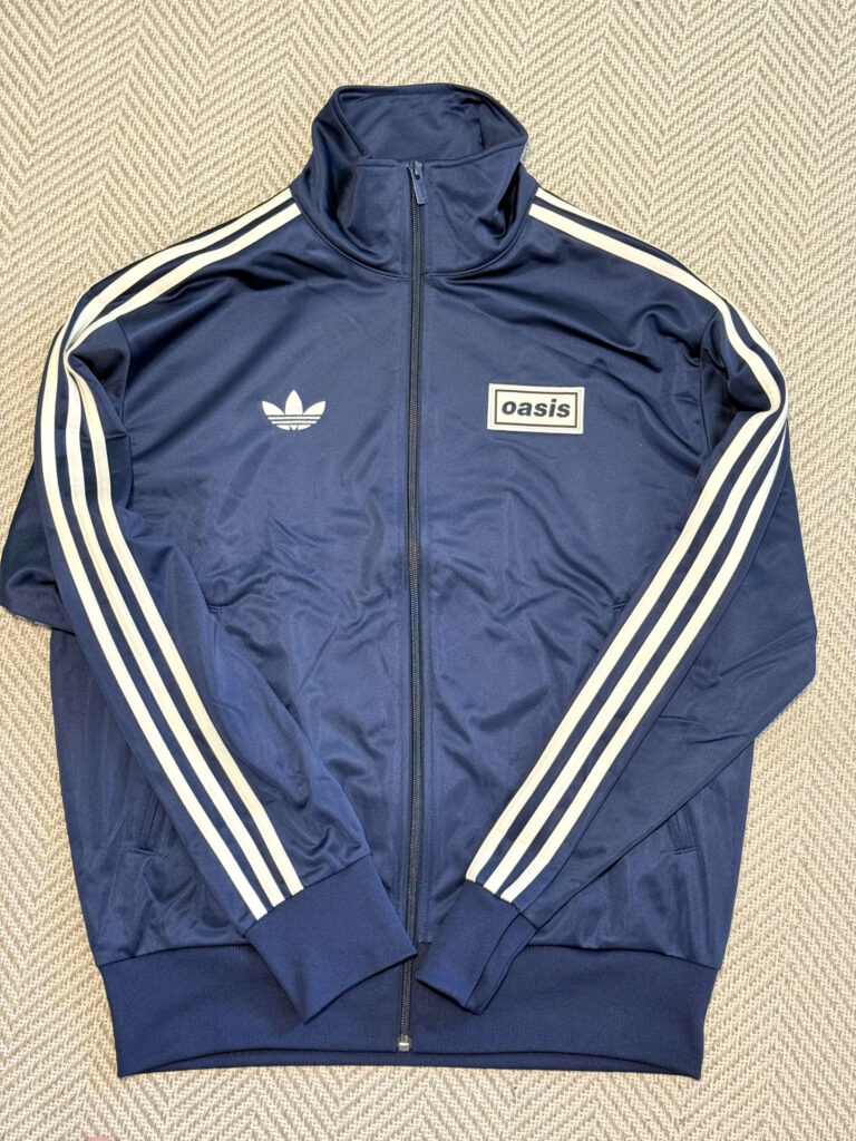 A navy blue Adidas track jacket with an Oasis logo laid flat on a beige herringbone-patterned surface. The jacket features the classic three white stripes on the sleeves.